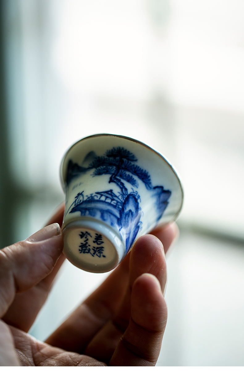 Close-up of a hand holding a porcelain teacup, showing the hand-painted landscape and silver trim.